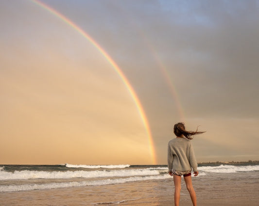 Woman facing away from camera and towards a double rainbow over the sea on a beach.
